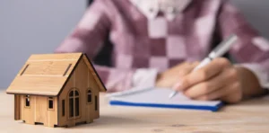 Person making a list of assets and a wooden model home on the table.
