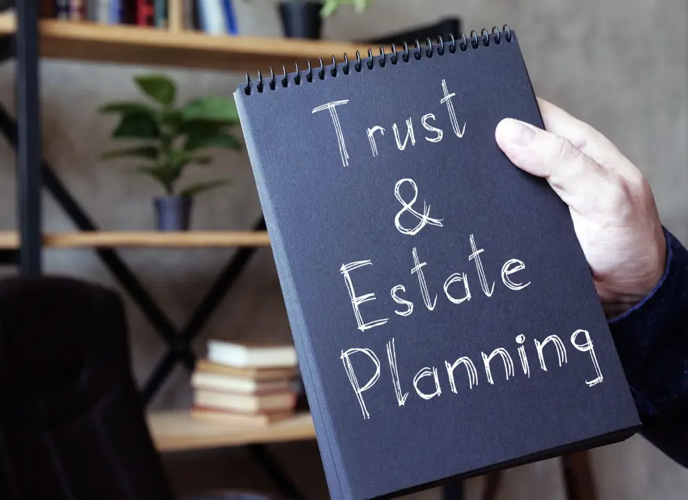 A person holding a black notebook labeled “Trust & Estate Planning” in a home office setting.