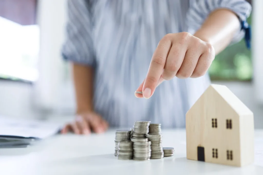 A woman stacking coins beside a miniature house, symbolizing estate or executor decisions.